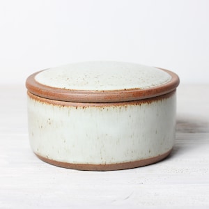 Op de afbeelding: Een witte keramische pot met een bruine rand en deksel. De pot heeft een gespikkeld glazuur en is perfect voor het bewaren van kleine voorwerpen.