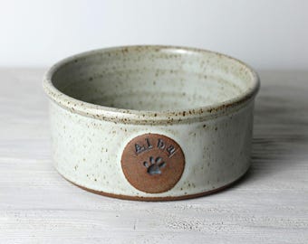 May include: A speckled, off-white ceramic pet bowl with a brown rim and base. A circular brown emblem with the name "ALBA" and a paw print is affixed to the bowl's side. The bowl is designed for pet food or water.