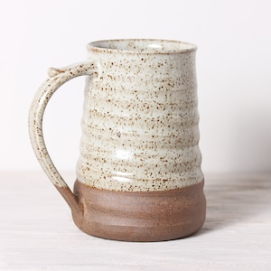 May include: A white ceramic mug with brown speckles and a brown rim. The mug has a wide, rounded handle.