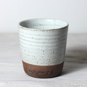 Pode incluir: Uma caneca de cerâmica artesanal com esmalte branco salpicado e base marrom. A palavra "Gramps" está inscrita na base. A caneca tem formato cilíndrico e aparência rústica e texturizada.