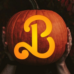Puede incluir: Una calabaza tallada con una gran letra naranja "B" en la parte delantera. La calabaza está siendo sostenida por las manos de una persona.