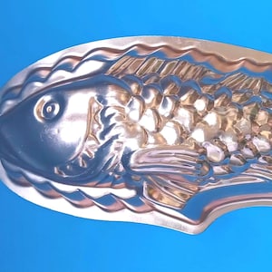 Puede incluir: Un molde de metal color cobre con forma de pez que presenta escamas, aletas y un ojo detallados en relieve. El molde tiene un borde festoneado e incluye un pequeño lazo de metal para colgar en el lado izquierdo y un asa de alambre en el lado derecho.