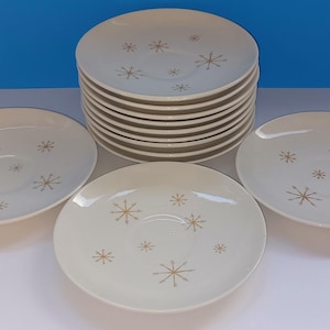 Puede incluir: Un conjunto de platillos de color crema con un diseño de estrellas doradas. Los platillos están apilados y dispuestos con tres platillos individuales. Cada platillo tiene un acabado liso y brillante. El fondo es azul claro.