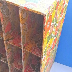K&ouml;nnte beinhalten: Ein vertikaler Aufbewahrungsorganizer aus Pappe mit einem sich wiederholenden Blumenmuster in Gelb-, Orange-, Rosa- und Gr&uuml;nt&ouml;nen. Die Einheit ist in sechs rechteckige F&auml;cher unterteilt, die in zwei Spalten und drei Reihen mit offenen Fronten angeordnet sind.
