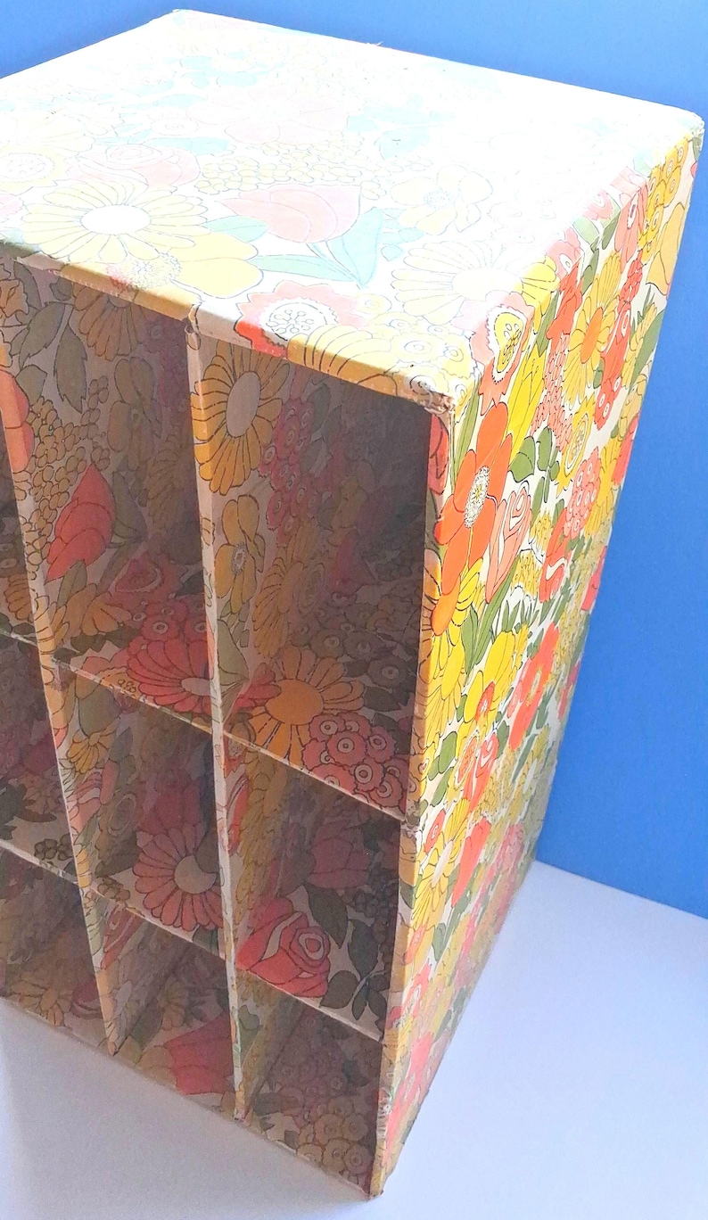 K&ouml;nnte beinhalten: Ein vertikaler Aufbewahrungsorganizer aus Pappe mit einem sich wiederholenden Blumenmuster in Gelb-, Orange-, Rosa- und Gr&uuml;nt&ouml;nen. Die Einheit ist in sechs rechteckige F&auml;cher unterteilt, die in zwei Spalten und drei Reihen mit offenen Fronten angeordnet sind.
