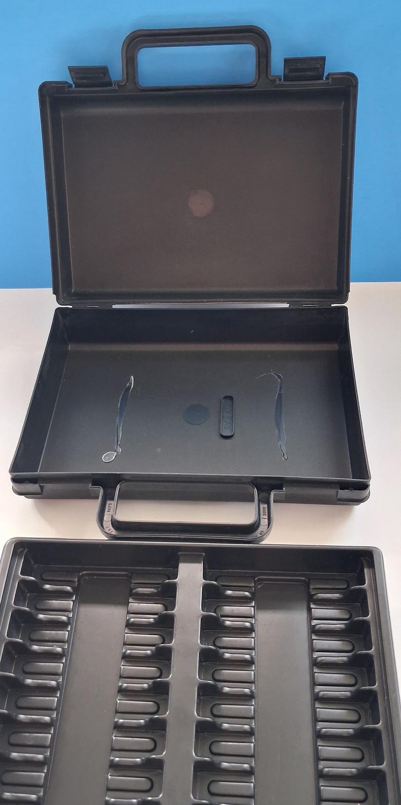 May include: Black plastic carrying case with an open lid and a separate tray. The case has a handle and a latch. The tray has multiple compartments. The background is white and blue.
