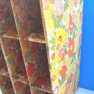 K&ouml;nnte beinhalten: Ein vertikaler Aufbewahrungsorganizer aus Pappe mit einem sich wiederholenden Blumenmuster in Gelb-, Orange-, Rosa- und Gr&uuml;nt&ouml;nen. Die Einheit ist in sechs rechteckige F&auml;cher unterteilt, die in zwei Spalten angeordnet sind, mit offenen Fronten und einer gl&auml;nzenden Oberfl&auml;che.
