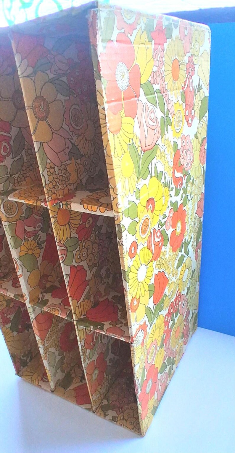 K&ouml;nnte beinhalten: Ein vertikaler Aufbewahrungsorganizer aus Pappe mit einem sich wiederholenden Blumenmuster in Gelb-, Orange-, Rosa- und Gr&uuml;nt&ouml;nen. Die Einheit ist in sechs rechteckige F&auml;cher unterteilt, die in zwei Spalten angeordnet sind, mit offenen Fronten und einer gl&auml;nzenden Oberfl&auml;che.