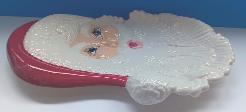 May include: A ceramic Santa Claus face-shaped dish. The dish features a red hat, a white beard, and a pink nose. The eyes are blue, and the dish is white. This decorative piece is perfect for holiday decor.