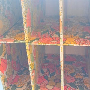 K&ouml;nnte beinhalten: Ein vertikaler Aufbewahrungsorganizer mit einer Gitterstruktur aus mehreren F&auml;chern. Die Innenfl&auml;chen sind mit einem sich wiederholenden Blumenmuster in leuchtenden Orange-, Gelb-, Rosa- und Gr&uuml;nt&ouml;nen auf cremefarbenem Grund versehen.