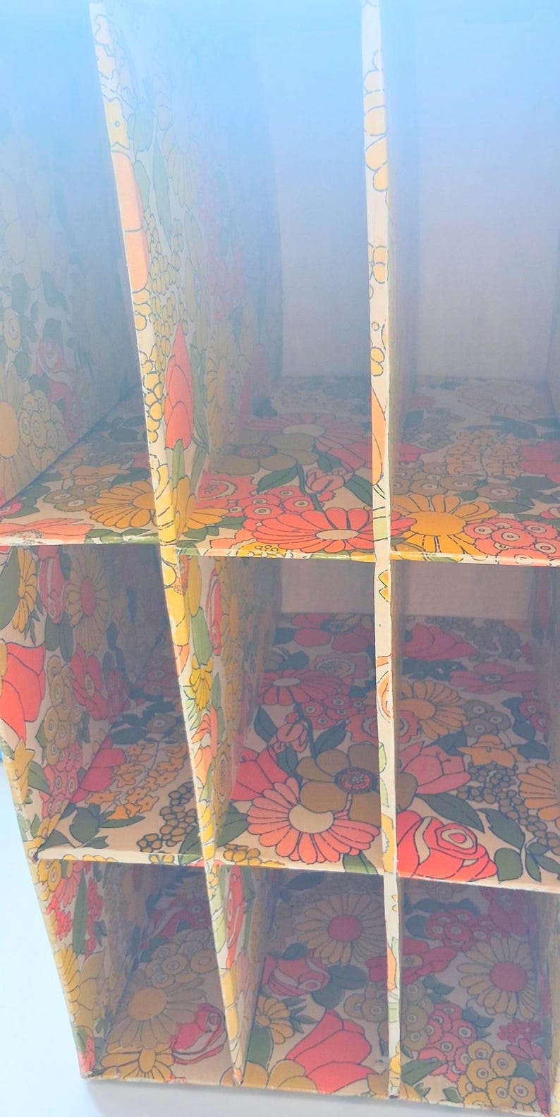 K&ouml;nnte beinhalten: Ein vertikaler Aufbewahrungsorganizer mit einer Gitterstruktur aus mehreren F&auml;chern. Die Innenfl&auml;chen sind mit einem sich wiederholenden Blumenmuster in leuchtenden Orange-, Gelb-, Rosa- und Gr&uuml;nt&ouml;nen auf cremefarbenem Grund versehen.
