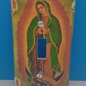 May include: A decorative light switch cover featuring an image of Our Lady of Guadalupe. The artwork depicts the Virgin Mary in a green robe with gold accents, set against a warm, radiant background. The cover has a blue switch opening and is secured with two screws.
