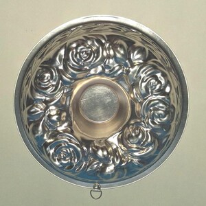 Puede incluir: Un molde de horneado redondo de metal color plata con un patr&oacute;n floral en relieve de rosas y hojas en las paredes interiores. El centro del molde es liso y circular, y tiene una peque&ntilde;a anilla de metal para colgar en el borde exterior.