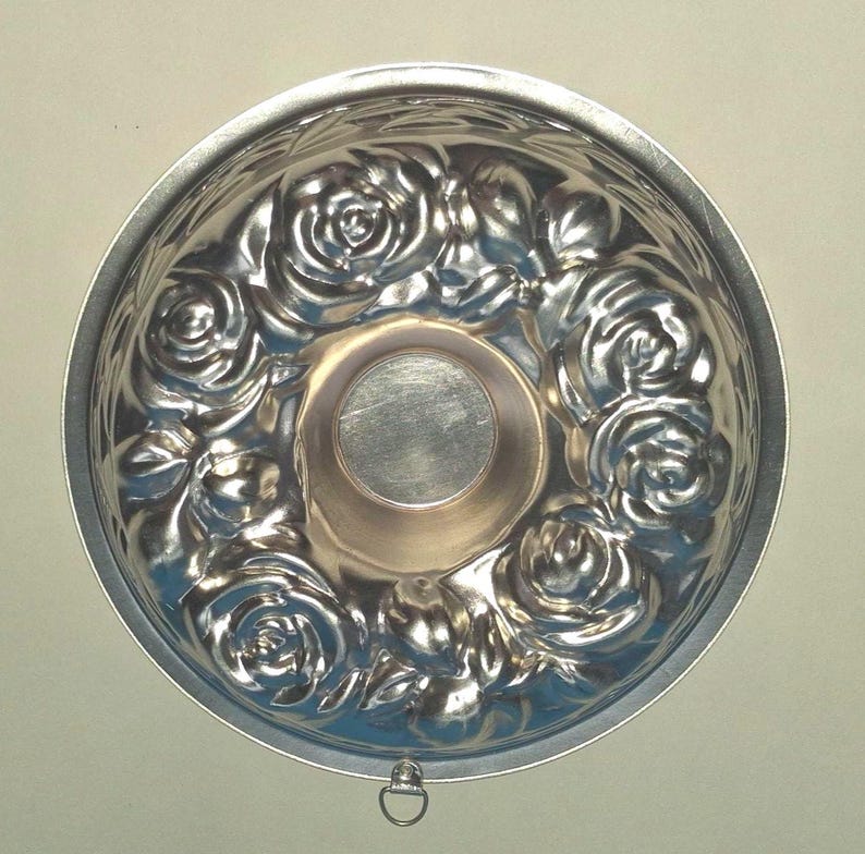 Puede incluir: Un molde de horneado redondo de metal color plata con un patr&oacute;n floral en relieve de rosas y hojas en las paredes interiores. El centro del molde es liso y circular, y tiene una peque&ntilde;a anilla de metal para colgar en el borde exterior.