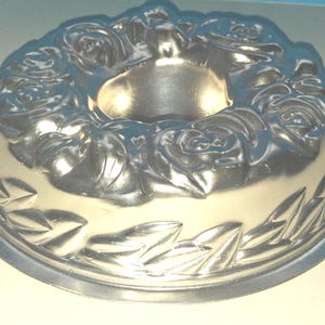 Puede incluir: Un molde para pastel tipo bundt de metal color plateado con un patr&oacute;n floral en relieve de rosas alrededor del borde superior y un borde decorativo de hojas en la parte inferior, con un tubo central hueco para hornear.