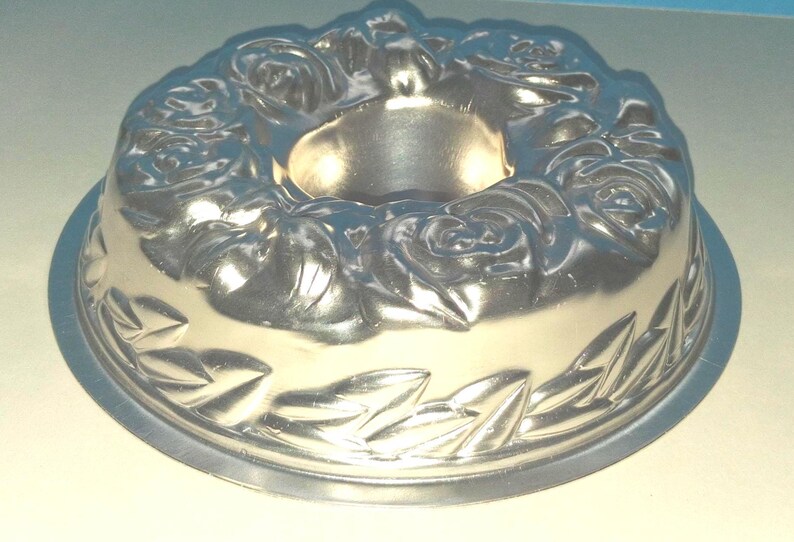 Puede incluir: Un molde para pastel tipo bundt de metal color plateado con un patr&oacute;n floral en relieve de rosas alrededor del borde superior y un borde decorativo de hojas en la parte inferior, con un tubo central hueco para hornear.