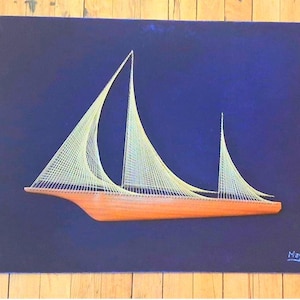 May include: A string art wall piece featuring a wooden sailboat hull base with light-colored thread creating the sails against a dark blue rectangular background. The name Mayes is written in white in the bottom right corner of the dark blue field.