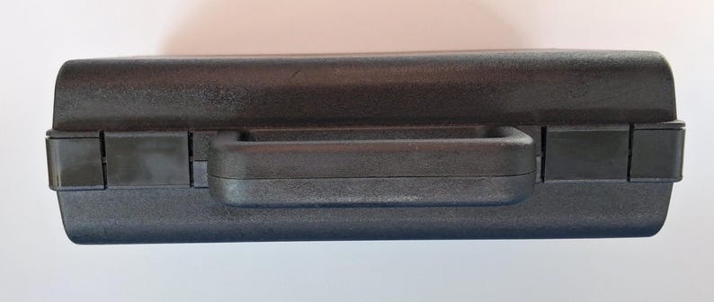 May include: A black, rectangular carrying case with a handle and two latches. The case is made of a textured material and appears to be closed. The handle is centered on the top of the case.