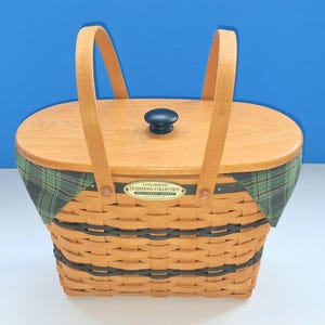 May include: A woven wooden basket with two upright handles and a removable wooden lid featuring a dark knob. The basket has a green and navy plaid fabric liner and a brass-colored plaque reading Longaberger Traditions Collection Fellowship Basket.