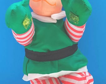 May include: A plush elf doll with a green tunic, hat, and shoes. The doll has a white collar, red and white striped pants, and green gloves with yellow accents. The elf's face is peach-colored with a small nose and orange mustache.