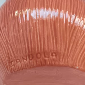 May include: Close-up of a ceramic rooster figurine in shades of orange, red, and yellow. The base of the figurine has the word "ANGOLA" imprinted. The rooster has a textured, ridged surface.