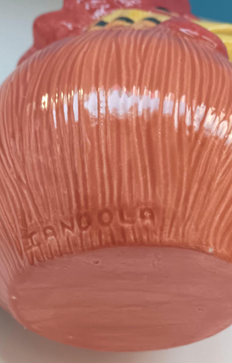 May include: Close-up of a ceramic rooster figurine in shades of orange, red, and yellow. The base of the figurine has the word "ANGOLA" imprinted. The rooster has a textured, ridged surface.