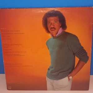 May include: Album cover featuring a portrait of Lionel Richie in a green sweater and pink collared shirt against an orange background. The album's tracklist is visible on the left side. The album is vintage.