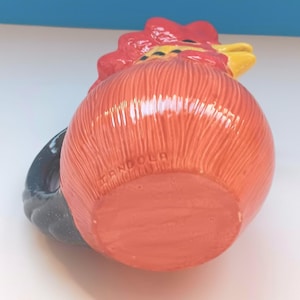 May include: A ceramic rooster-shaped mug with a red comb, orange body, and black handle. The mug has a textured surface and the word "ANDOLA" is printed on the bottom. The rooster's beak is yellow.