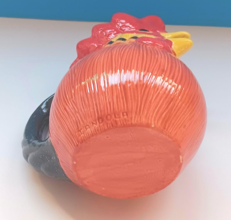 May include: A ceramic rooster-shaped mug with a red comb, orange body, and black handle. The mug has a textured surface and the word "ANDOLA" is printed on the bottom. The rooster's beak is yellow.