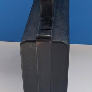 May include: A black, rectangular carrying case with a handle on top. The case has a textured surface and a secure latch. The handle is integrated into the top of the case, providing a comfortable grip.