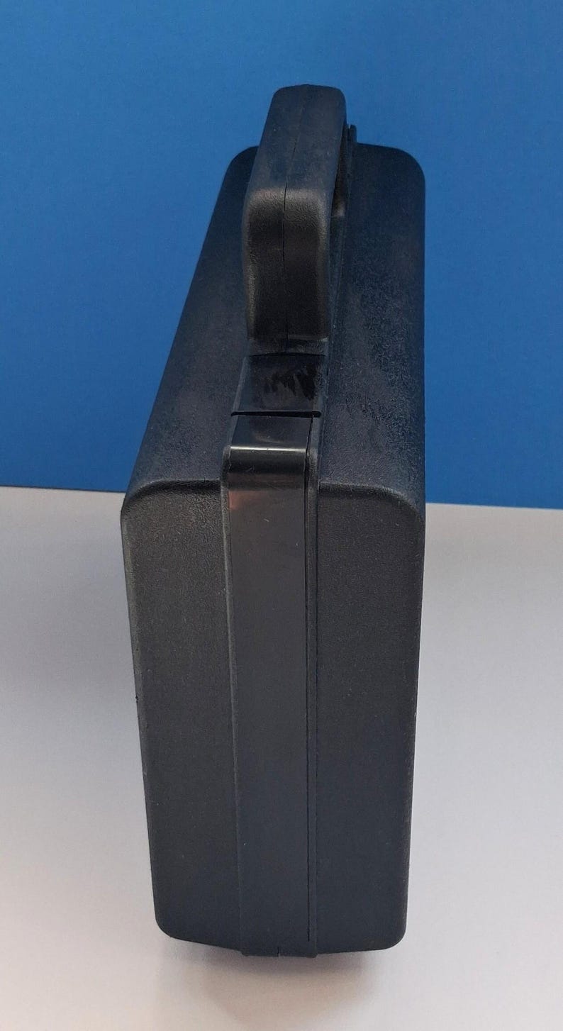 May include: A black, rectangular carrying case with a handle on top. The case has a textured surface and a secure latch. The handle is integrated into the top of the case, providing a comfortable grip.