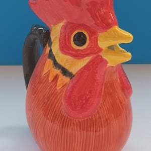 May include: A ceramic pitcher shaped like a rooster, featuring a red comb, wattle, and body. The pitcher has a black handle and yellow beak. The design is detailed with textured lines, creating a rustic, farmhouse aesthetic.