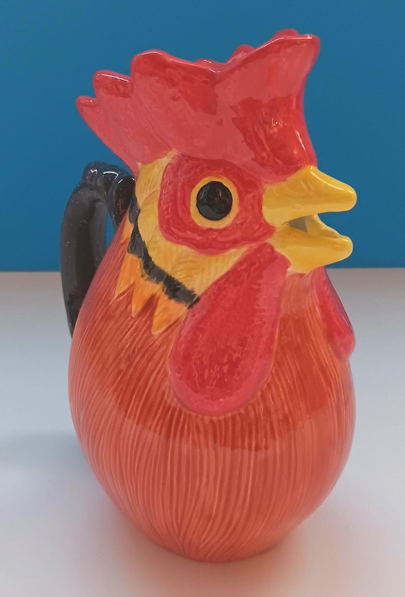 May include: A ceramic pitcher shaped like a rooster, featuring a red comb, wattle, and body. The pitcher has a black handle and yellow beak. The design is detailed with textured lines, creating a rustic, farmhouse aesthetic.