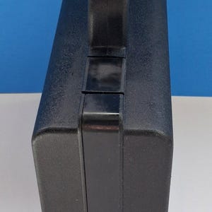 May include: A black, rectangular carrying case with a textured surface and a sturdy handle. The case is designed for portability and protection, suitable for tools or equipment. The handle is securely attached to the top.