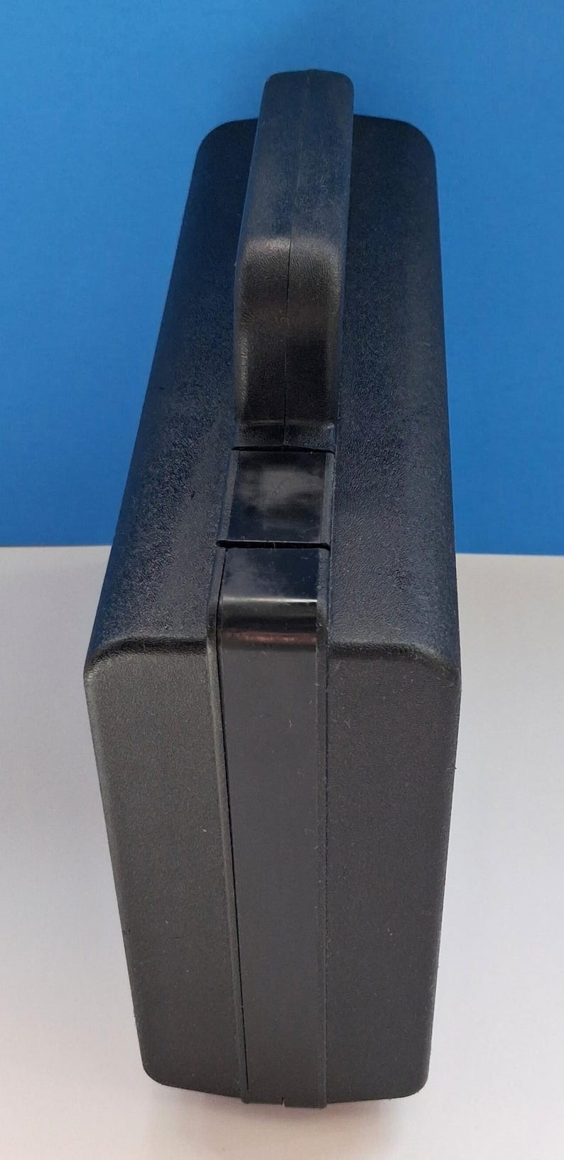 May include: A black, rectangular carrying case with a textured surface and a sturdy handle. The case is designed for portability and protection, suitable for tools or equipment. The handle is securely attached to the top.