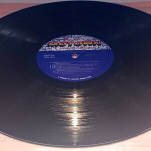 May include: A black vinyl record with a blue and white label featuring the Motown logo. The record is on a light pink background with text. The label lists song titles and artist names. The record is a vintage music item.
