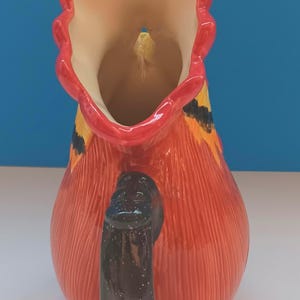 May include: A ceramic pitcher with a red body and a black handle. The pitcher has a decorative design with yellow and orange accents. The rim is red and the interior is cream-colored. The pitcher is likely used for serving beverages.