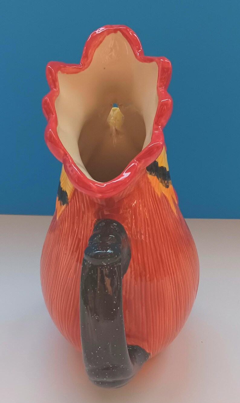 May include: A ceramic pitcher with a red body and a black handle. The pitcher has a decorative design with yellow and orange accents. The rim is red and the interior is cream-colored. The pitcher is likely used for serving beverages.