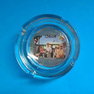 May include: A glass ashtray with a silver glitter surface and a picture of the band AC/DC on the front. The band members are standing in front of a building with the band's name in large letters above them.