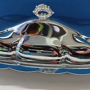 May include: A silver butter dish with a scalloped edge and a lid with a decorative knob. The dish is made of polished metal and reflects light.