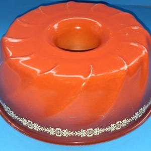 Puede incluir: Molde para bizcocho naranja y marrón con un patrón floral blanco en la base. El molde tiene un acabado brillante y una forma circular con un agujero en el centro, ideal para hornear pasteles.