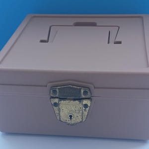 May include: A tan, rectangular box with a handle on the lid and a metal clasp. The clasp is gold-colored and has the words "Safety Mint" on it. The box is closed and appears to be made of plastic.