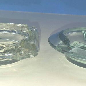 May include: Two clear glass ashtrays, one square with rounded corners and one circular. The square ashtray features a visible embossed text on the bottom that reads OPORTI A800. Both items have molded notches along the edges for resting items.