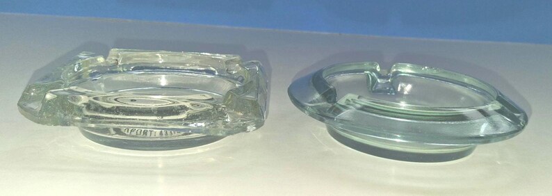 May include: Two clear glass ashtrays, one square with rounded corners and one circular. The square ashtray features a visible embossed text on the bottom that reads OPORTI A800. Both items have molded notches along the edges for resting items.