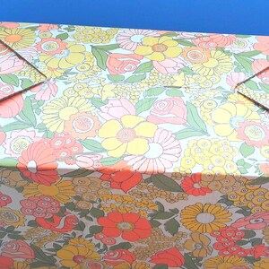 K&ouml;nnte beinhalten: Ein rechteckiger Pappkarton mit einem dichten, sich wiederholenden Blumenmuster in den Farben Orange, Gelb, Rosa und Gr&uuml;n. Der Karton hat einen gefalteten Deckel mit vier dreieckigen Laschen, die in der Mitte zusammenlaufen.