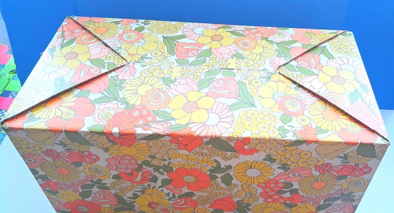 K&ouml;nnte beinhalten: Ein rechteckiger Pappkarton mit einem dichten, sich wiederholenden Blumenmuster in den Farben Orange, Gelb, Rosa und Gr&uuml;n. Der Karton hat einen gefalteten Deckel mit vier dreieckigen Laschen, die in der Mitte zusammenlaufen.