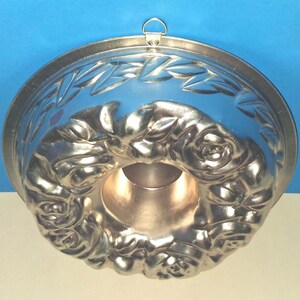 Puede incluir: Un molde de reposter&iacute;a redondo de metal plateado con un patr&oacute;n de rosas en relieve alrededor del tubo central y un motivo de hojas en el borde exterior. Una peque&ntilde;a anilla met&aacute;lica para colgar est&aacute; fijada en el borde superior.