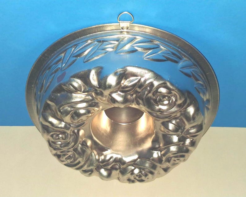 Puede incluir: Un molde de reposter&iacute;a redondo de metal plateado con un patr&oacute;n de rosas en relieve alrededor del tubo central y un motivo de hojas en el borde exterior. Una peque&ntilde;a anilla met&aacute;lica para colgar est&aacute; fijada en el borde superior.