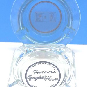 May include: A clear glass ashtray with a square base and a circular top rim. The center features a circular logo with the text Fontana's Spaghetti House, Cheektowaga: 822-7918, and Lockport: 433-4446. The upper section displays a faint R+A logo.