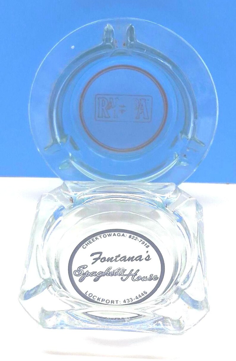 May include: A clear glass ashtray with a square base and a circular top rim. The center features a circular logo with the text Fontana's Spaghetti House, Cheektowaga: 822-7918, and Lockport: 433-4446. The upper section displays a faint R+A logo.